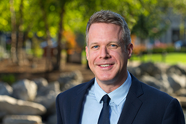 Headshot of employee Barrett Coleman in a navy blazer in park with rocks, water and green trees.